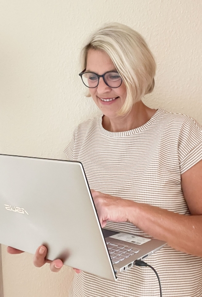 Patricia Wandt- Bürodienstleistungen - steht lächelnd mit einem Laptop an der Wand